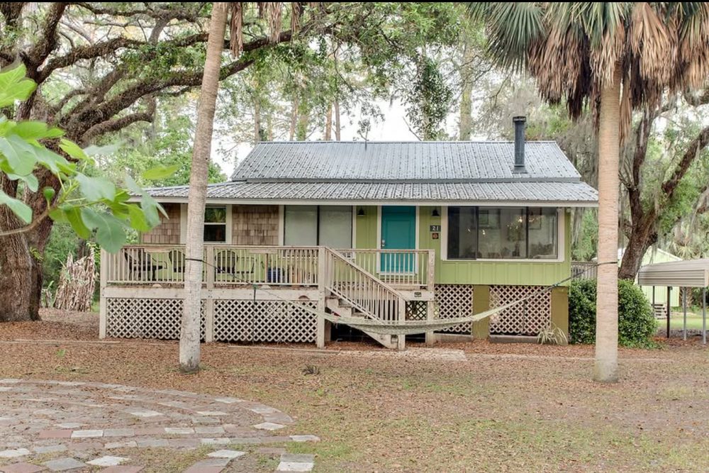 Green raised cottage with a front deck under oak trees